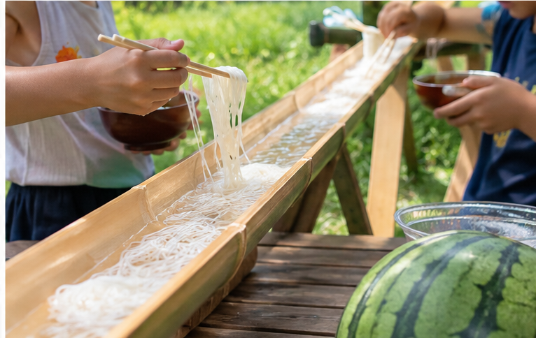 Family members catching somen noodles flowing through a bamboo channel outdoors.